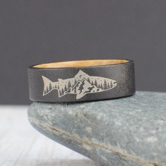 A sandblasted tungsten ring featuring an engraved fish with a mountain and tree design inside. The interior is lined with smooth, sanded whiskey barrel wood for a comfortable fit. This ring is perfect for everyday wear and makes a great gift for outdoor enthusiasts.