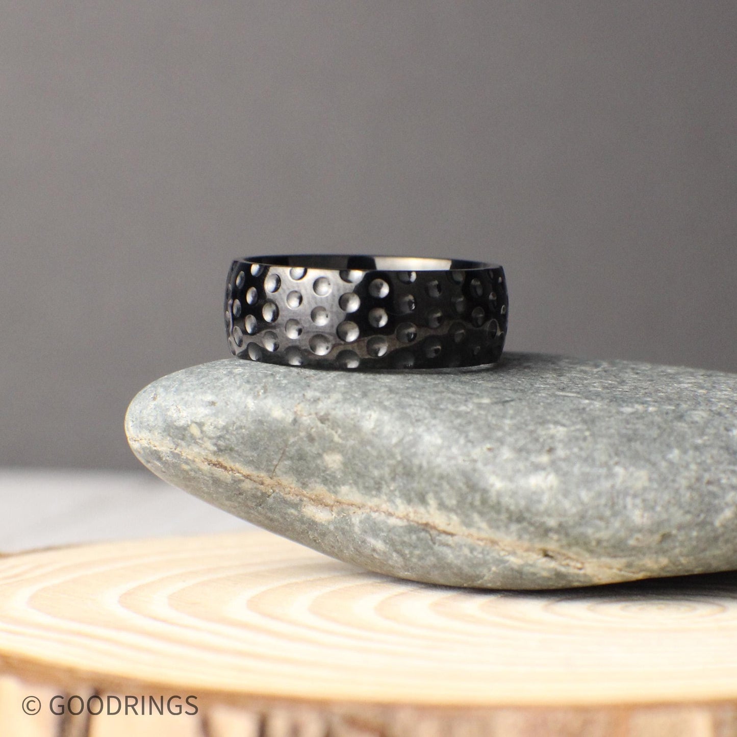 Unique 8mm wide titanium golf ball textured ring with a hammered-like dimple design across the surface, photographed in a rustic natural setting on stone and wood, perfect for couples looking for a durable and sporty wedding band or anniversary gift inspired by the game of golf.