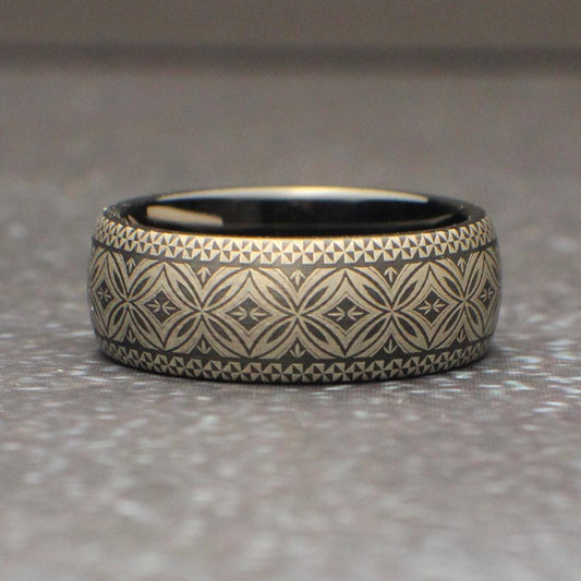 Close up of a black tungsten ring featuring a continuous Polynesian tribal engraving pattern wrapping around the entire band. The silver gray etched design contrasts against the dark metal creating a bold mens wedding band photographed on natural wood.