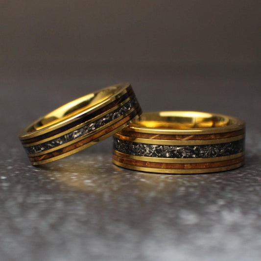 Picture of an 8mm wide gold ring with a 6mm wide gold ring leaning on it. The rings are made out of gold, tungsten, meteorite, and whiskey barrel wood.