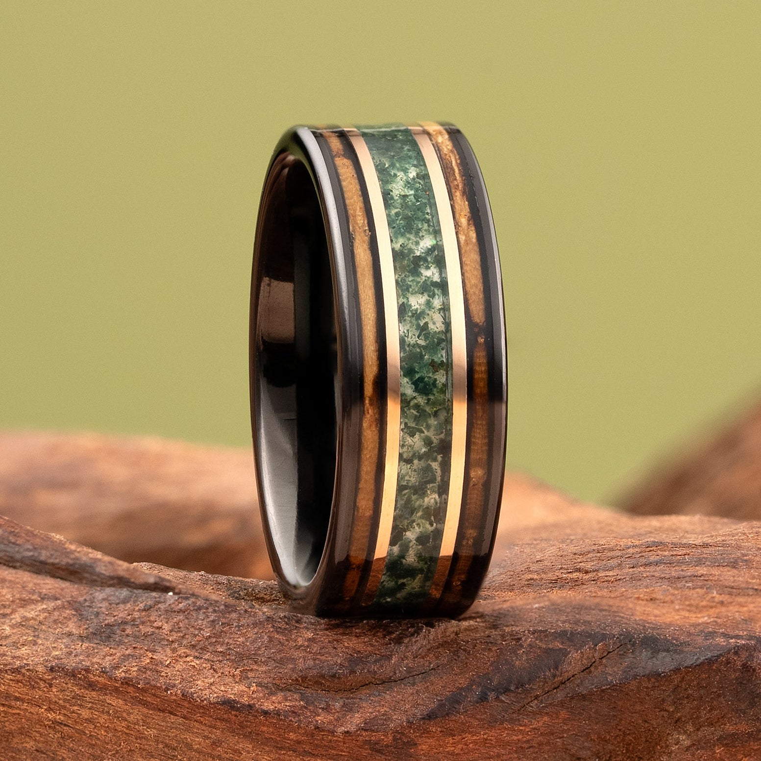 A black tungsten ring with a green and gold inlay, standing upright on a piece of natural driftwood against a solid olive-green background.
