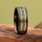 A black tungsten ring with a green and gold inlay, standing upright on a piece of natural driftwood against a solid olive-green background.