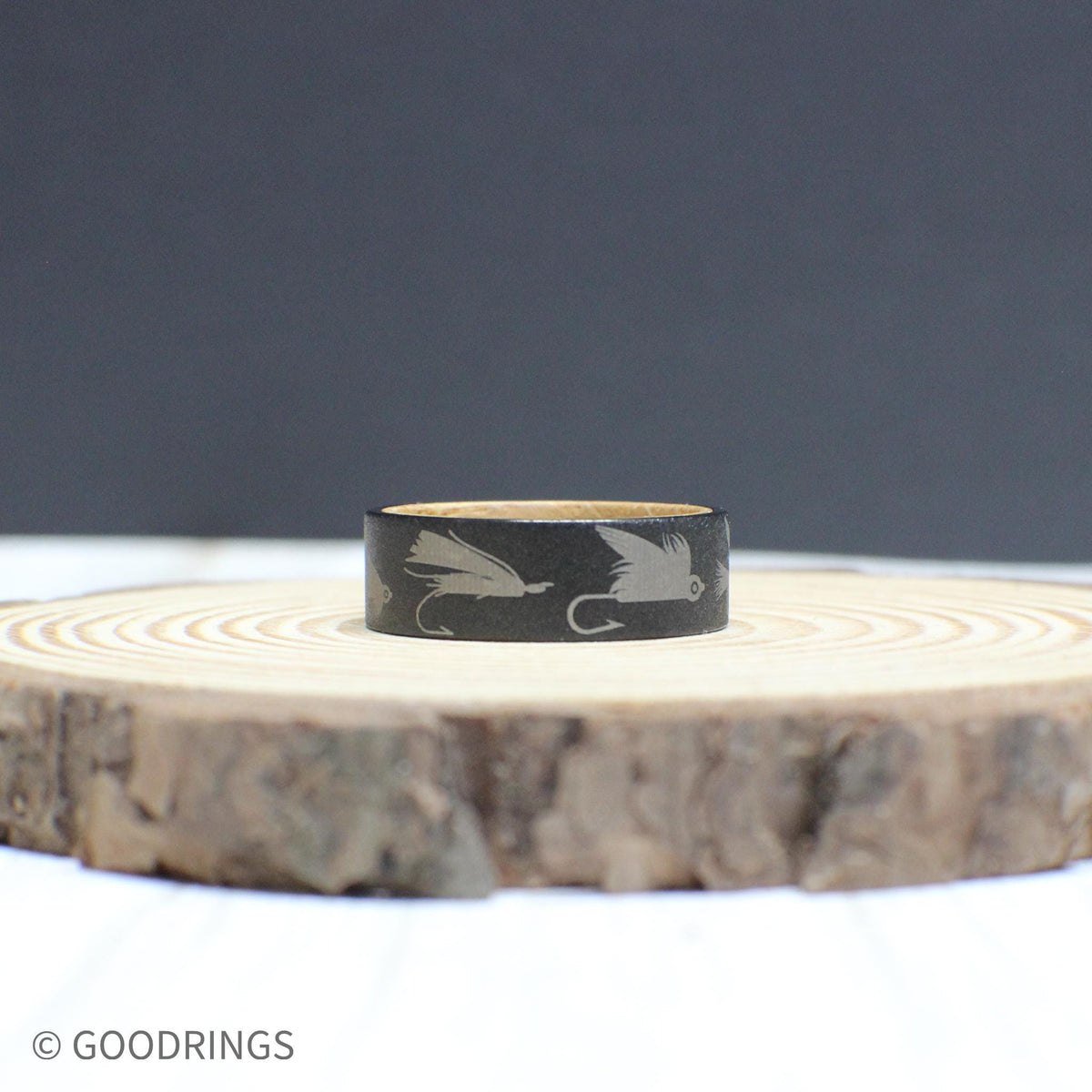 Fly fishing-themed tungsten ring with a sandblasted texture, engraved lure designs, and a whiskey barrel wood inlay.
