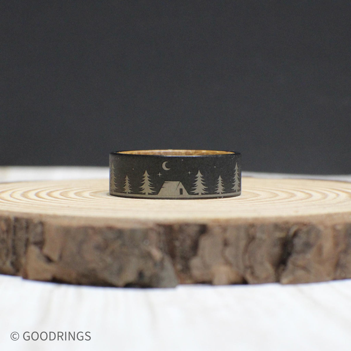 Tungsten ring showcasing an engraved outdoor scene with trees, a tent, and a bear under a starlit sky. The inside features warm whiskey barrel wood.