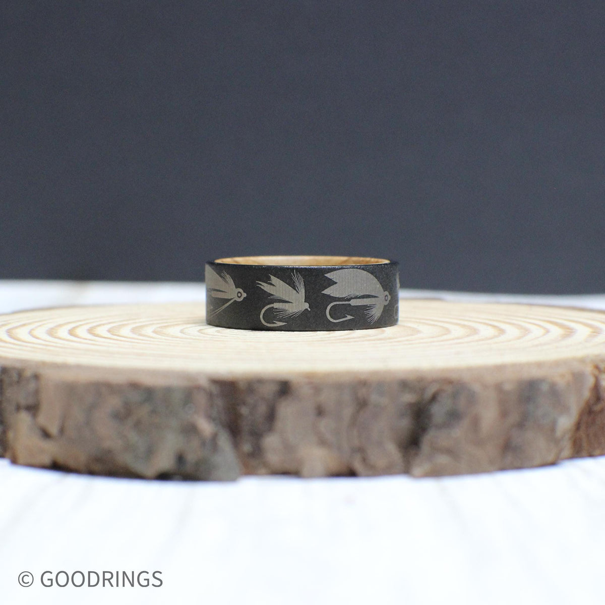 A unique fly fishing ring made from tungsten with engraved lure designs and a whiskey barrel wood interior.