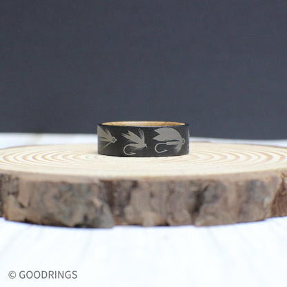 A unique fly fishing ring made from tungsten with engraved lure designs and a whiskey barrel wood interior.