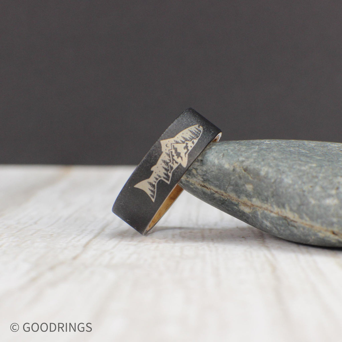 This tungsten ring features a fish engraving with a mountain and tree design inside, set on a sandblasted exterior. The inside is lined with smooth, sanded whiskey barrel wood for comfort, making it an ideal gift for outdoor enthusiasts.