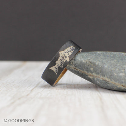 This tungsten ring features a fish engraving with a mountain and tree design inside, set on a sandblasted exterior. The inside is lined with smooth, sanded whiskey barrel wood for comfort, making it an ideal gift for outdoor enthusiasts.