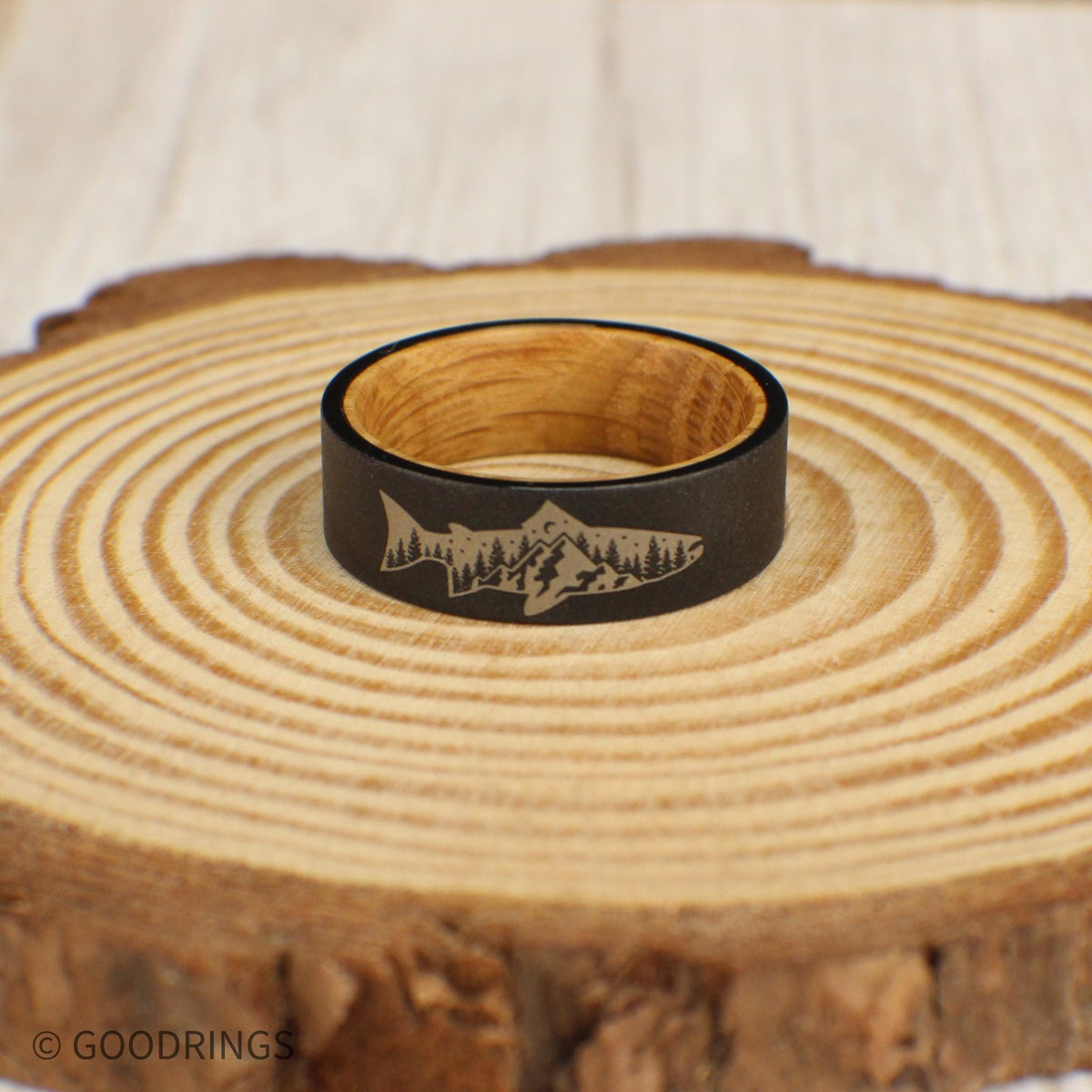 A unique tungsten ring featuring a sandblasted exterior with an engraved fish design. Inside the fish, there is a mountain and tree scene, and the interior is lined with sanded whiskey barrel wood for a comfortable, natural feel.