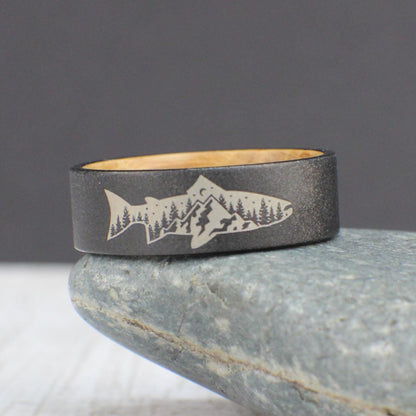 A sandblasted tungsten ring featuring an engraved fish with a mountain and tree design inside. The interior is lined with smooth, sanded whiskey barrel wood for a comfortable fit. This ring is perfect for everyday wear and makes a great gift for outdoor enthusiasts.