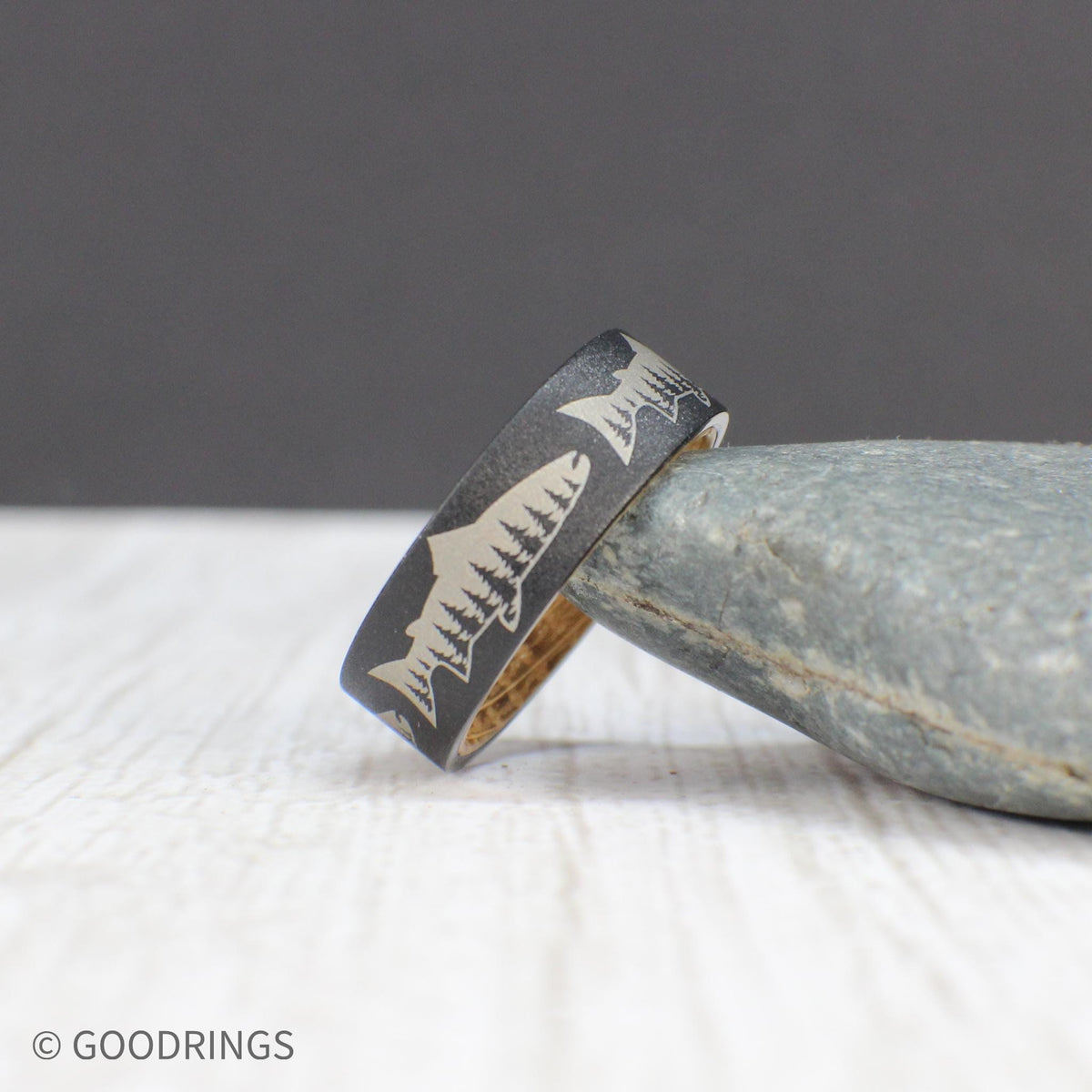 Close-up of a tungsten ring showcasing a salmon and tree line engraving, with a warm whiskey wood interior.