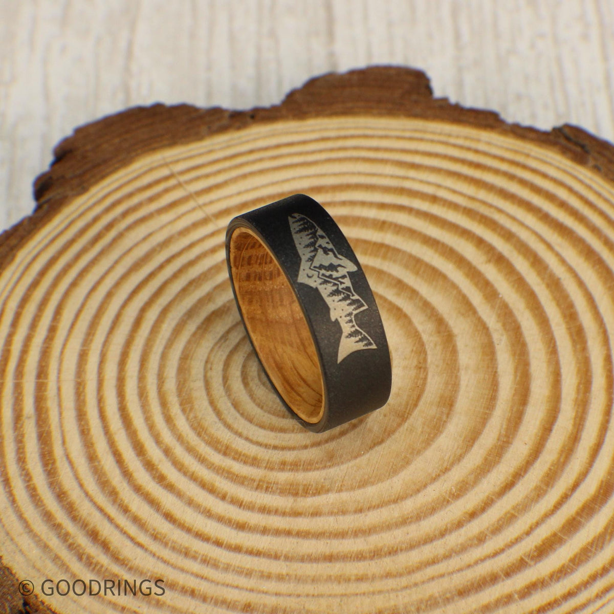 A sandblasted tungsten ring with an engraved fish design that includes a mountain and trees inside. The interior is lined with sanded whiskey barrel wood for a smooth, comfortable fit. This ring is a thoughtful gift for nature lovers.