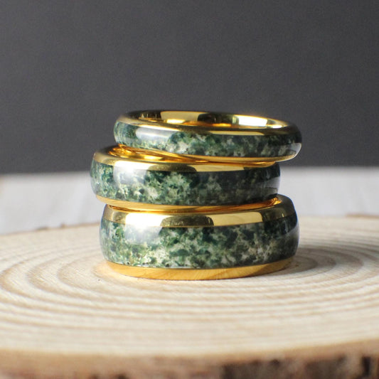 A handcrafted tungsten ring plated in yellow gold with a domed profile and an inlay of genuine moss agate. The natural green inclusions of the agate create a unique, organic pattern in every ring. The inlay is sealed with a crystal-clear resin to protect against moisture and wear. Available in 4mm, 6mm, and 8mm widths, this ring blends rugged durability with earthy elegance, making it ideal as a nature-inspired wedding band.