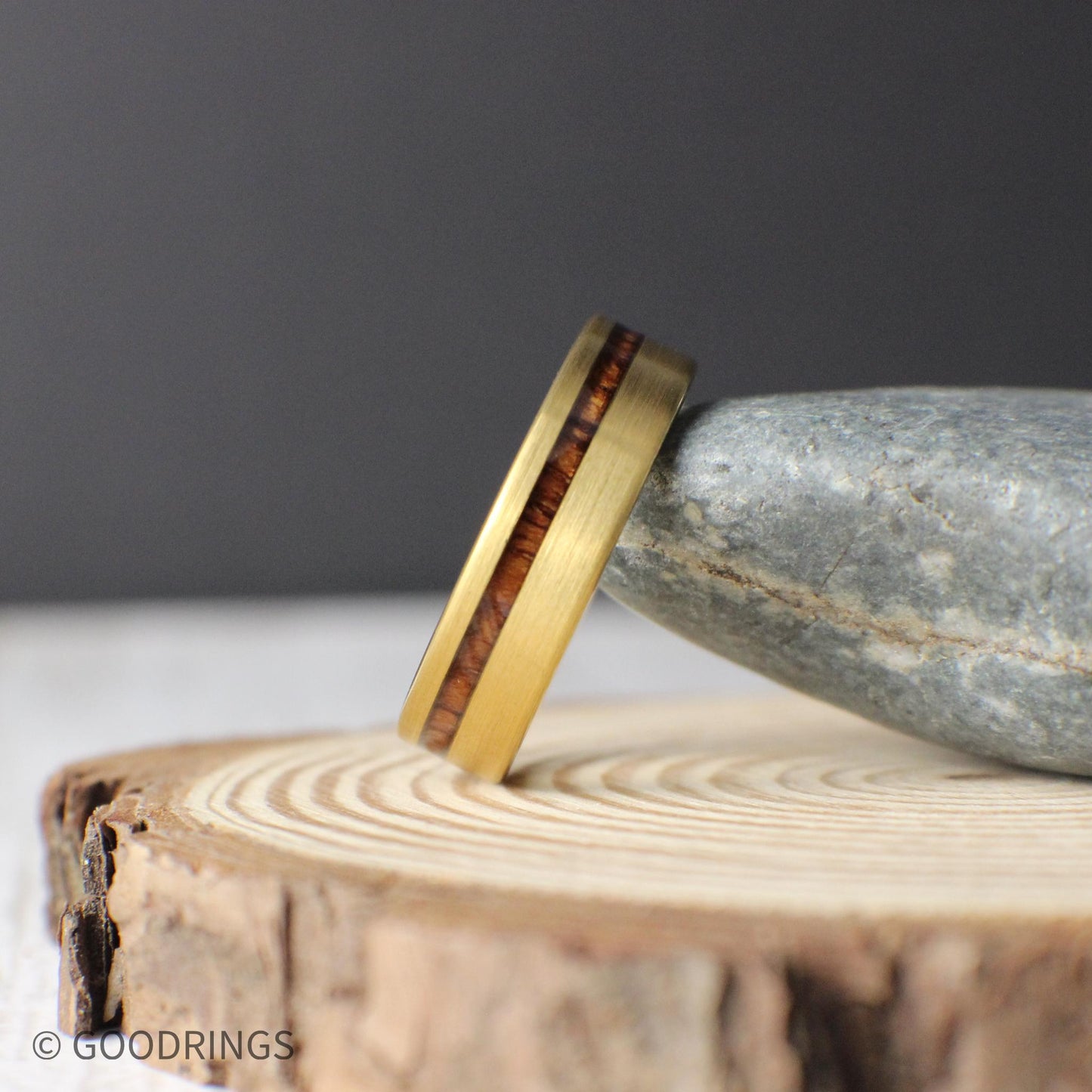 Close-up view of a gold tungsten ring with a warm Koa wood inlay running off-center. This modern men’s wedding band is crafted from tungsten carbide with a brushed gold finish, combining natural elements and industrial durability in one timeless piece.