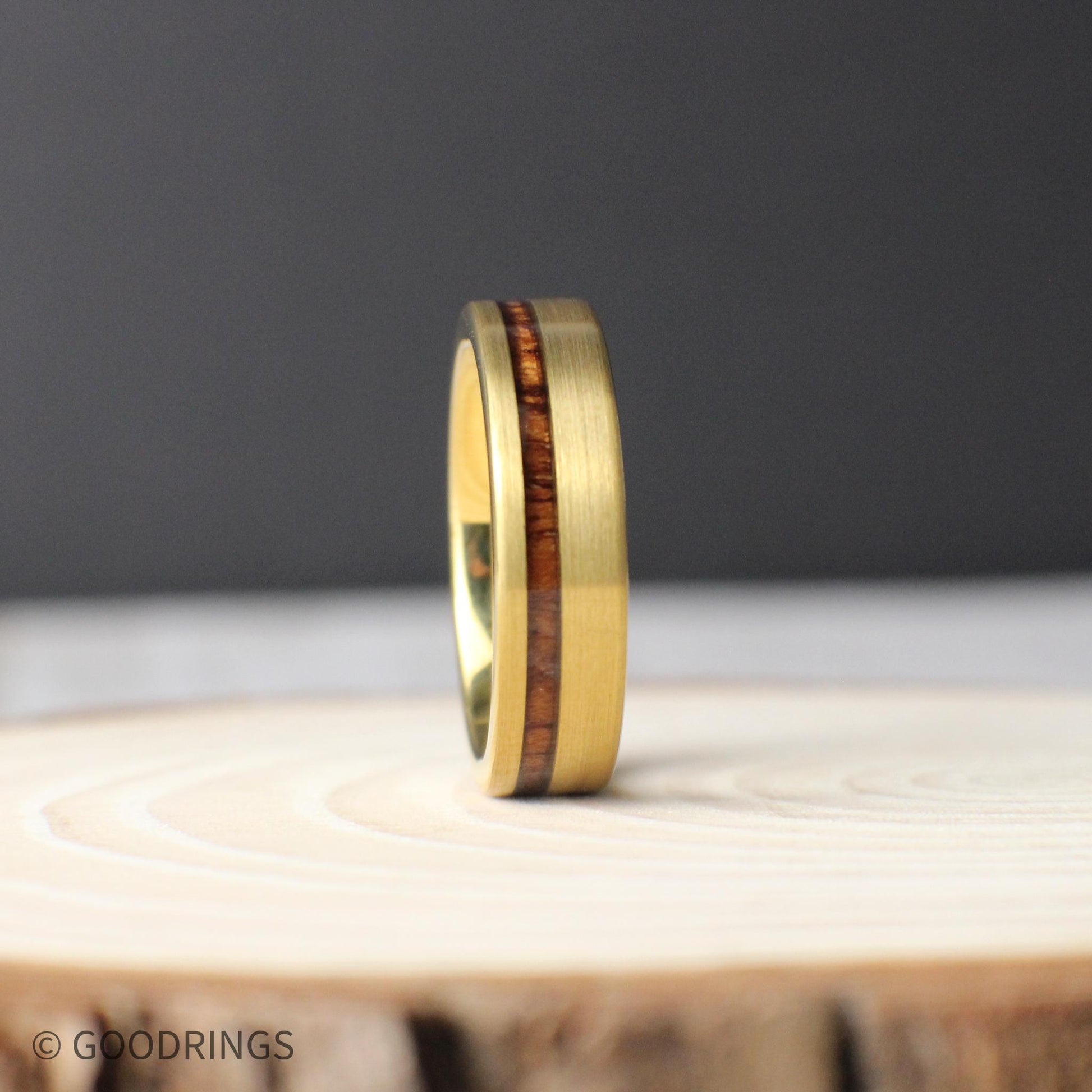 Standing upright on a wooden slab, this 6mm men’s tungsten ring showcases its offset inlay of Hawaiian Koa wood. The polished gold interior and brushed exterior reflect light subtly, while the unique grain of the Koa adds warmth and character.