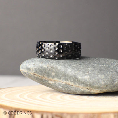 Unique 8mm wide titanium golf ball textured ring with a hammered-like dimple design across the surface, photographed in a rustic natural setting on stone and wood, perfect for couples looking for a durable and sporty wedding band or anniversary gift inspired by the game of golf.