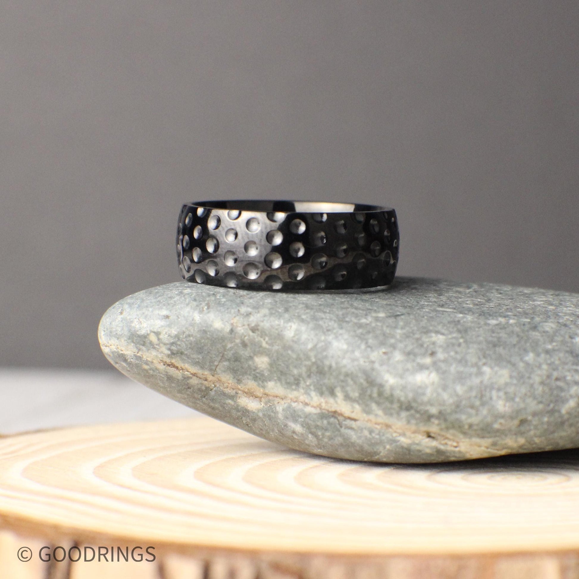 Unique 8mm wide titanium golf ball textured ring with a hammered-like dimple design across the surface, photographed in a rustic natural setting on stone and wood, perfect for couples looking for a durable and sporty wedding band or anniversary gift inspired by the game of golf.