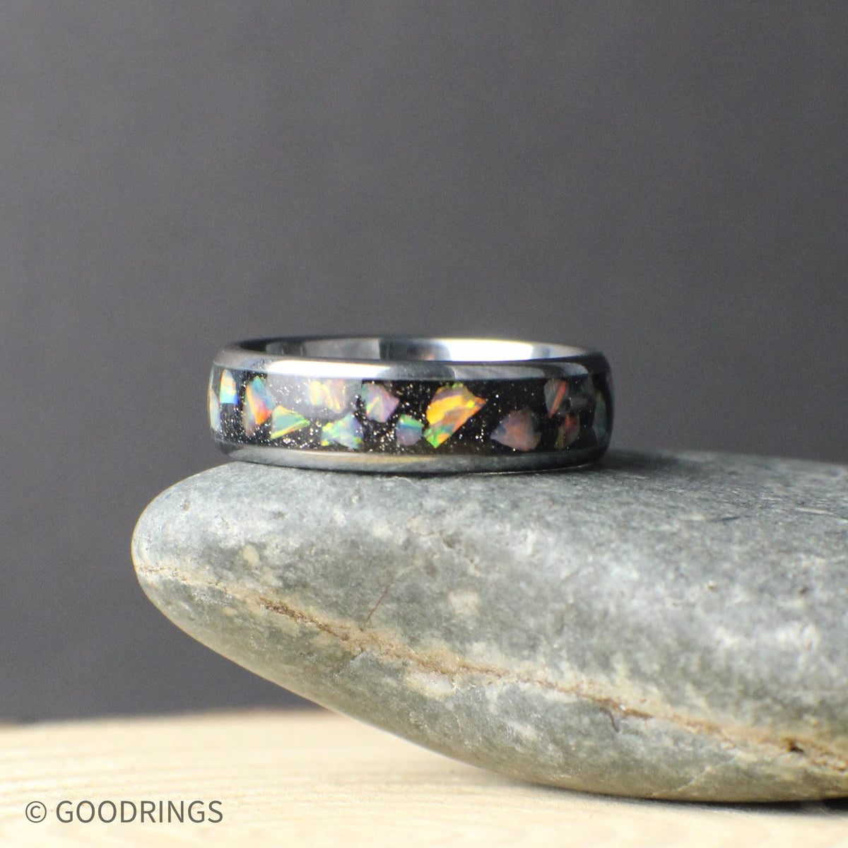 Top-down view of a polished tungsten opal band sitting on a stone surface. The crushed opal inlay runs continuously around the ring, showing natural variation in color and pattern. The slim 6mm width creates a clean, modern look suited for a wedding band, promise ring, or everyday wear.