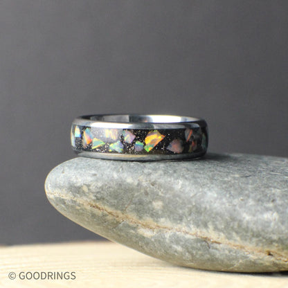 Top-down view of a polished tungsten opal band sitting on a stone surface. The crushed opal inlay runs continuously around the ring, showing natural variation in color and pattern. The slim 6mm width creates a clean, modern look suited for a wedding band, promise ring, or everyday wear.