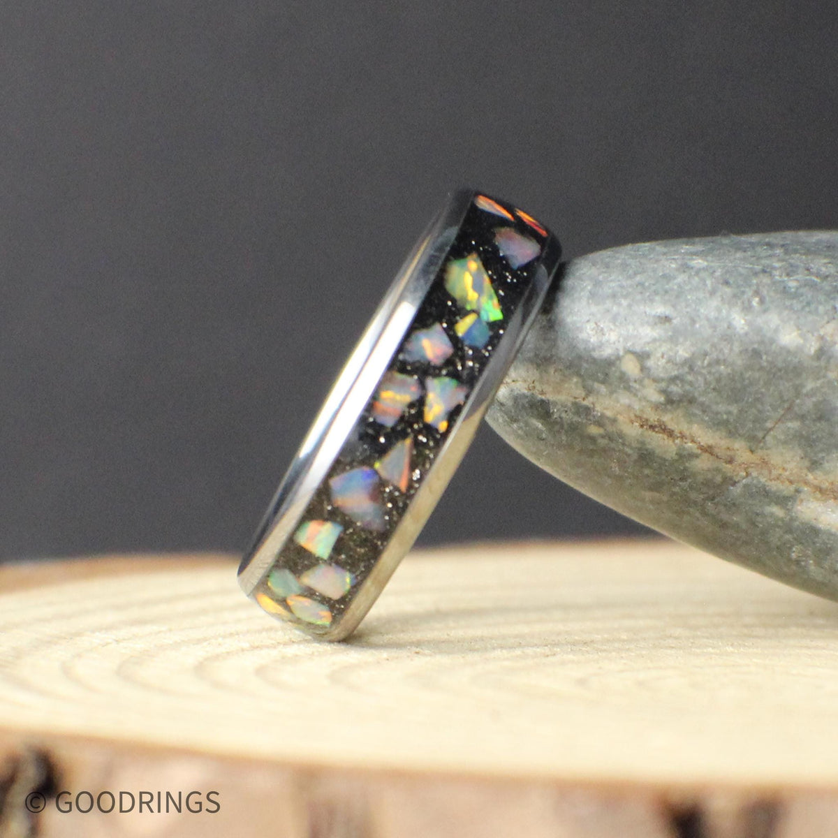 Side view of a narrow 6mm tungsten ring with a vibrant crushed opal inlay. The opal pieces reflect multicolor tones including green, teal, and gold, set into a dark tungsten base. The ring is photographed leaning against a smooth stone, emphasizing its durable build and refined, minimalist style.