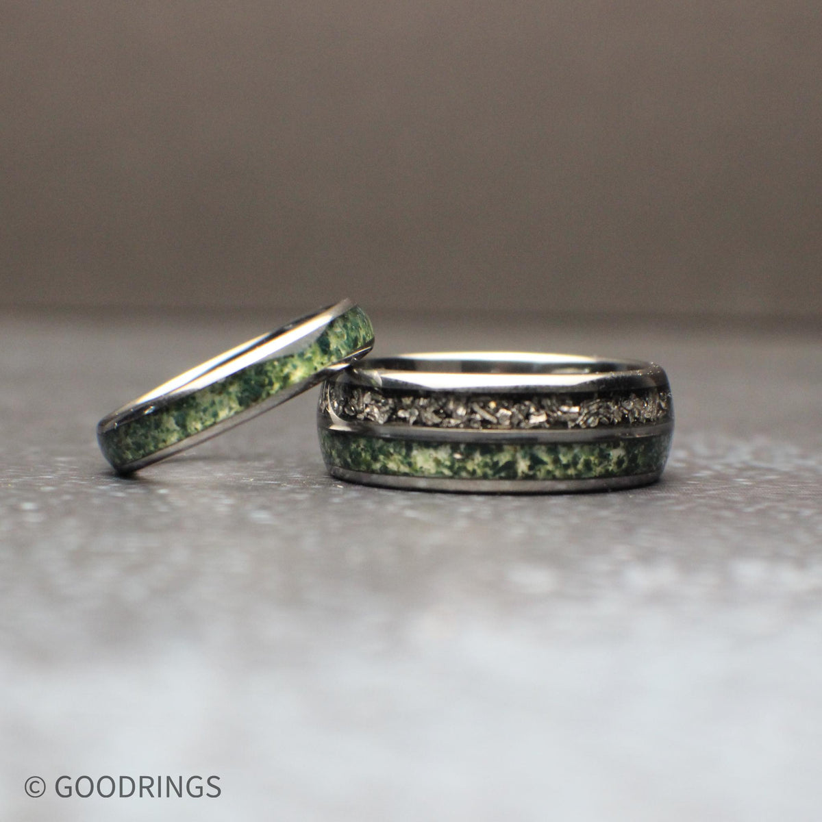 Couples wedding ring set featuring an 8mm black tungsten band with genuine meteorite and moss agate inlays, paired with a matching 4mm black ring featuring a moss agate inlay. The rings are displayed on a neutral textured surface, highlighting the natural green patterns of the moss agate and the unique texture of the meteorite for a modern, nature-inspired matching ring set.