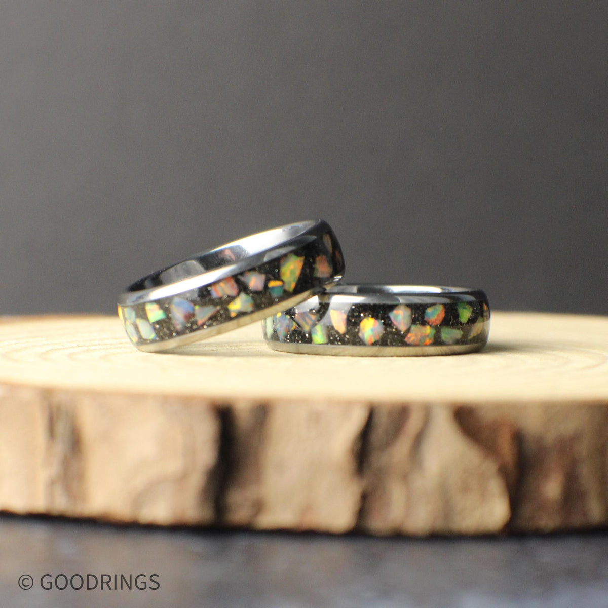 A tungsten carbide ring with a colorful opal inlay displayed on natural wood. The crushed opal catches the light with shifting colors, contrasting against the dark metal band. The smooth edges and narrow width give the ring a comfortable fit and a timeless appearance suitable for long-term wear.