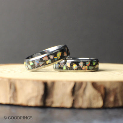 A tungsten carbide ring with a colorful opal inlay displayed on natural wood. The crushed opal catches the light with shifting colors, contrasting against the dark metal band. The smooth edges and narrow width give the ring a comfortable fit and a timeless appearance suitable for long-term wear.