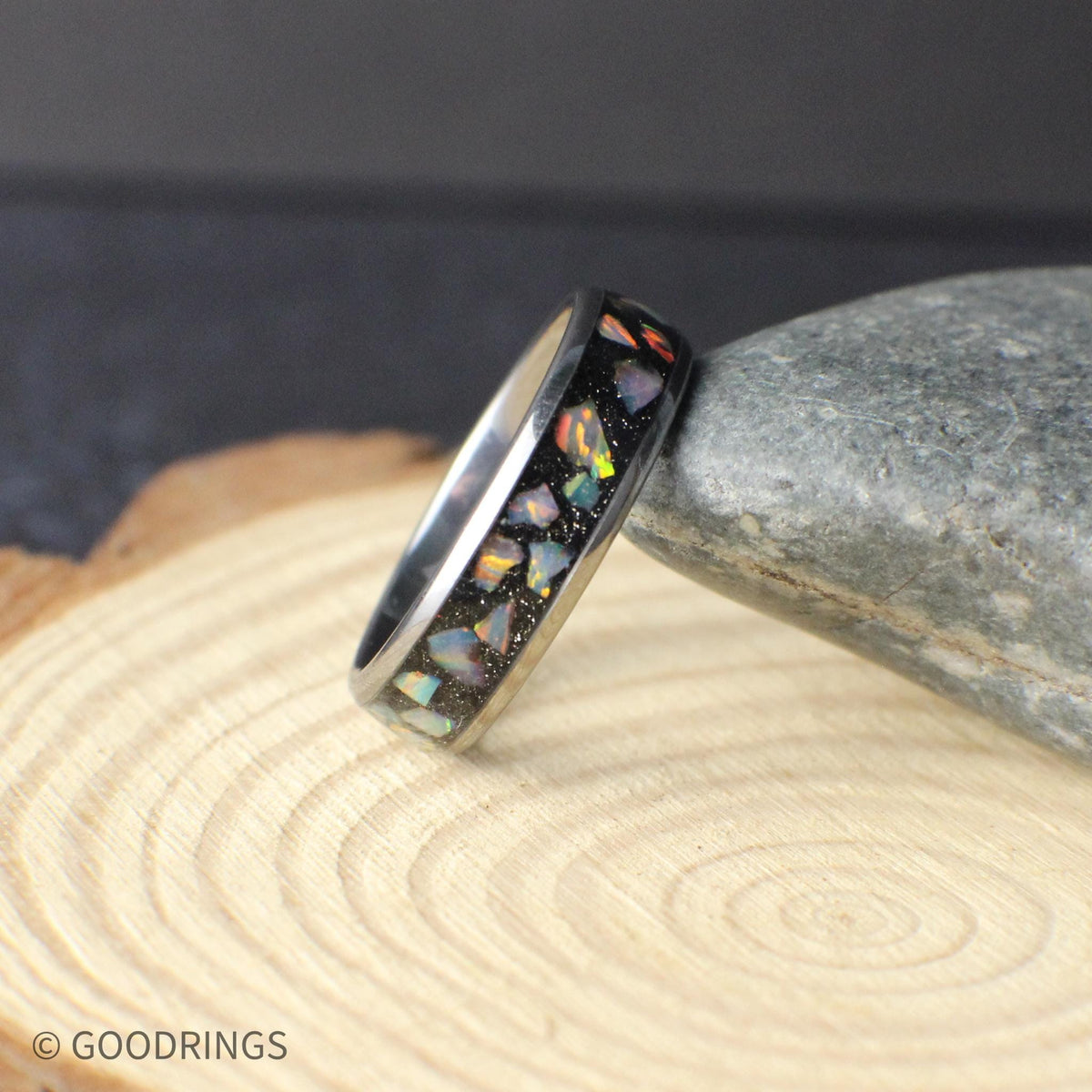 Minimalist tungsten opal wedding band photographed on a stone and wood surface. The polished 6mm band features a crushed opal inlay with natural color variation, offering a balance of durability and elegance. Designed for everyday wear, this ring combines modern materials with a timeless silhouette.