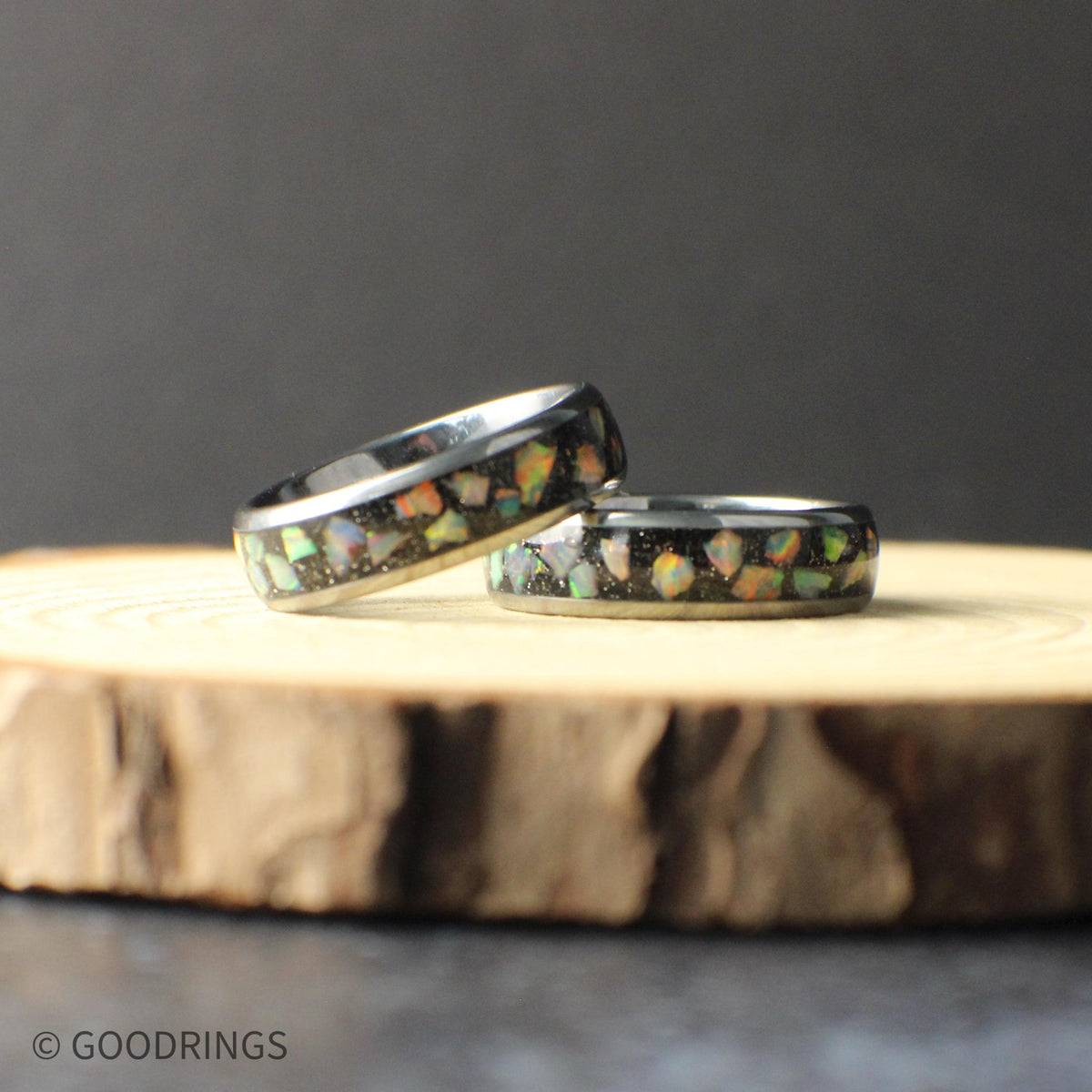 Close-up of a 6mm tungsten opal ring resting on a natural wood surface. The polished tungsten band features a crushed opal inlay with flashes of green, blue, and orange that shimmer against the dark background. The slim profile highlights the modern design, making it ideal for a wedding band or everyday ring with a subtle pop of color.
