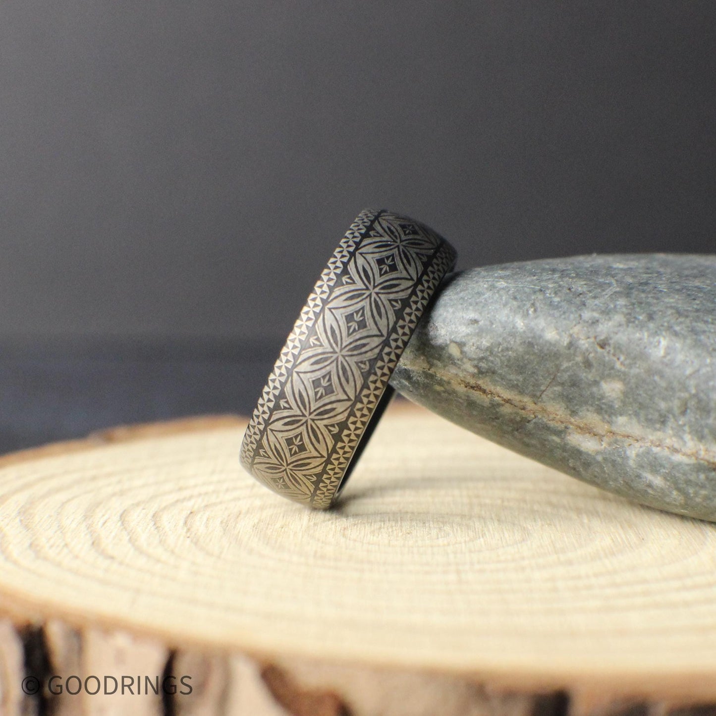 Angled product photo of a Polynesian style tungsten wedding band resting on a smooth stone. The engraved tribal pattern runs fully around the ring showing a detailed geometric motif symbolizing unity strength and protection.