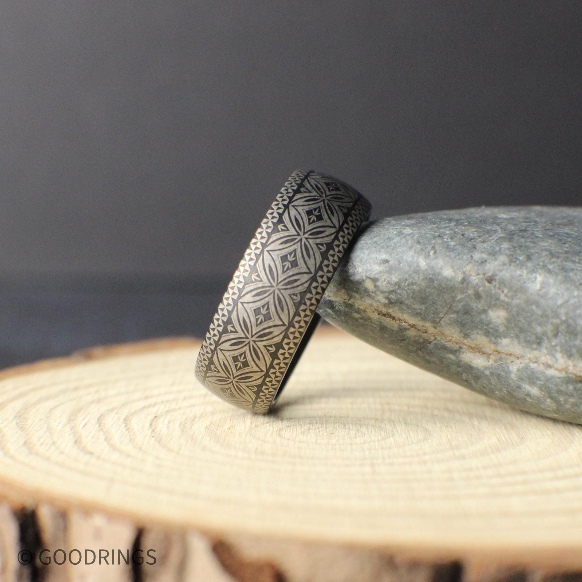 Angled product photo of a Polynesian style tungsten wedding band resting on a smooth stone. The engraved tribal pattern runs fully around the ring showing a detailed geometric motif symbolizing unity strength and protection.