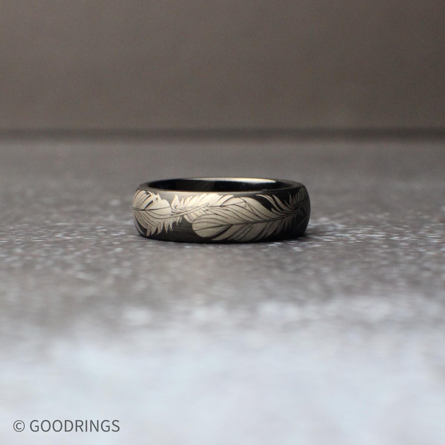 Detailed macro view of a 6mm comfort fit black tungsten band engraved with flowing feather artwork around the entire circumference of the ring.