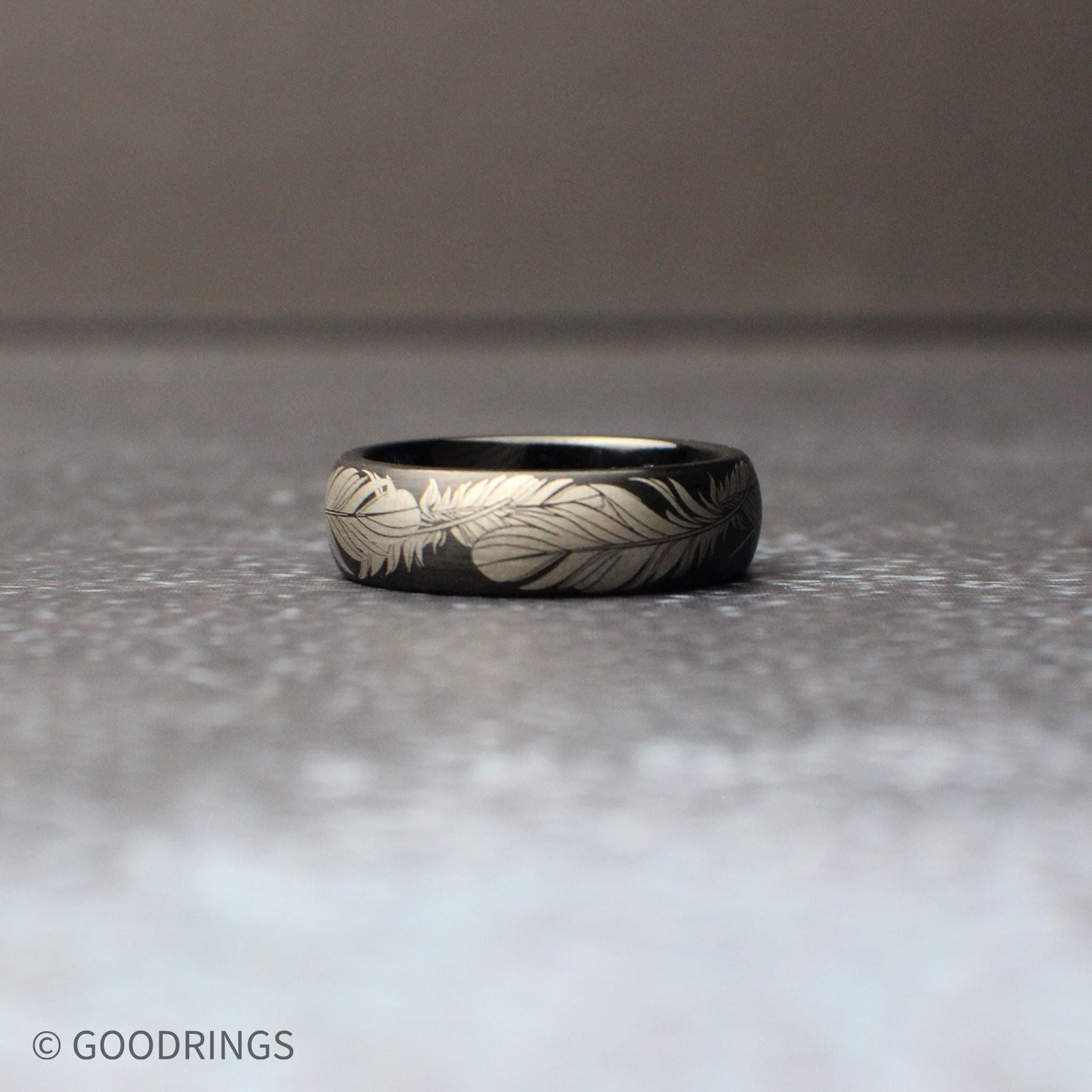 Detailed macro view of a 6mm comfort fit black tungsten band engraved with flowing feather artwork around the entire circumference of the ring.
