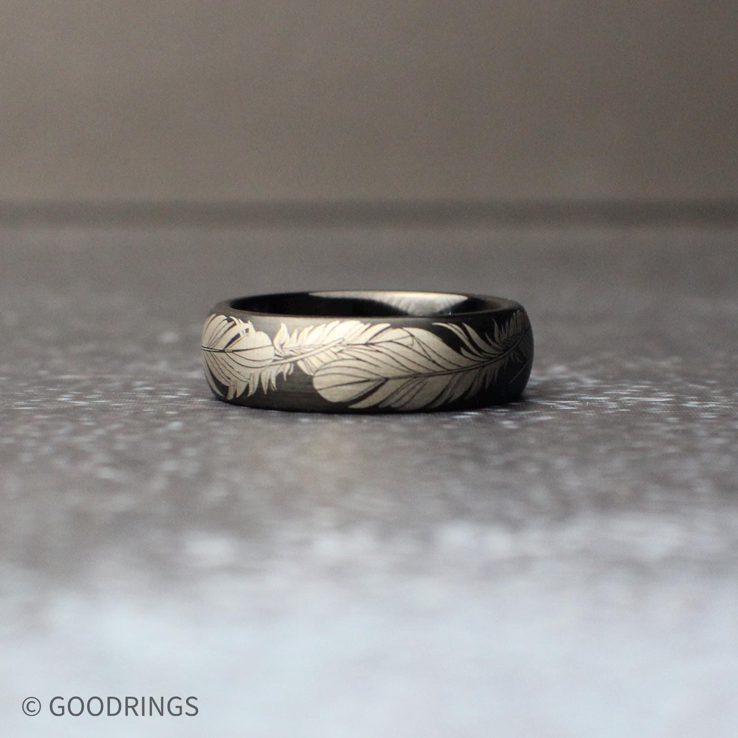 Nature-inspired 6mm black tungsten wedding band featuring an intricate feather design etched in silver tone, photographed in soft natural lighting.