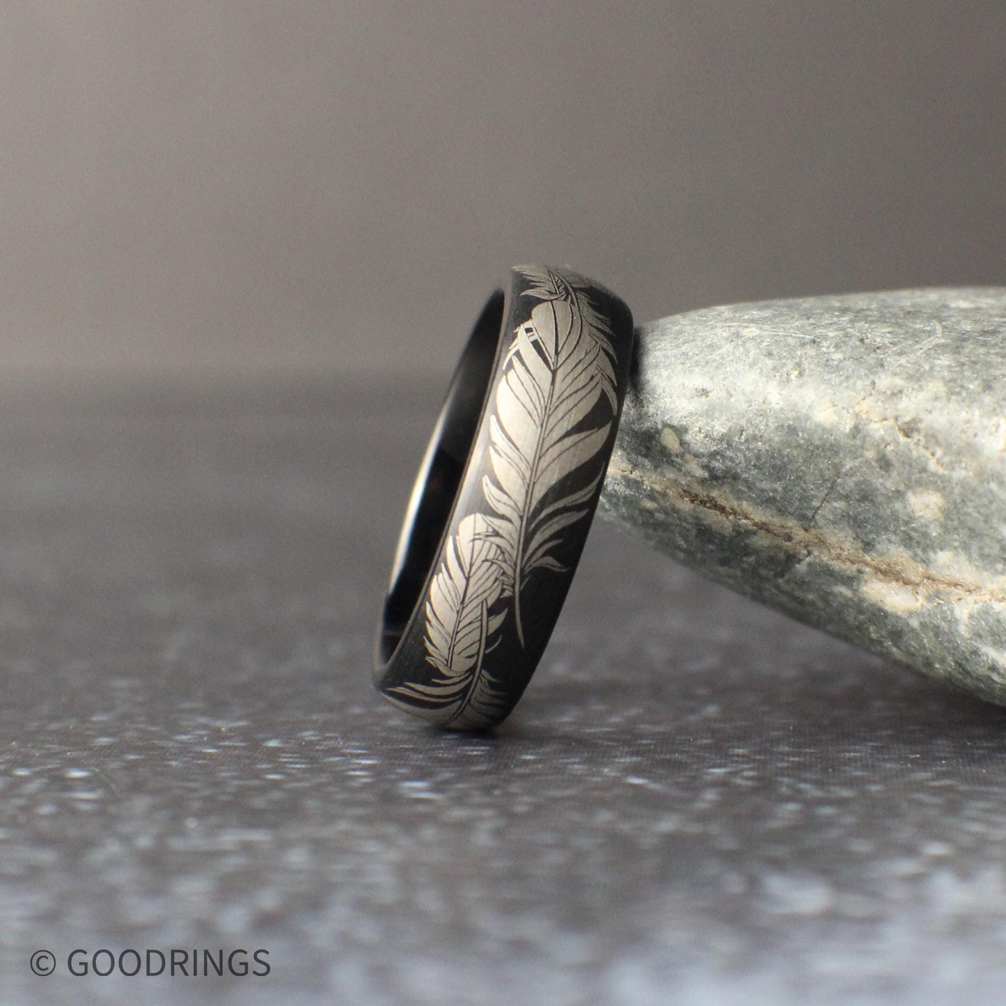 Men’s black tungsten ring with full wrap feather engraving, polished finish, and smooth rounded edges, displayed on a neutral gray backdrop.