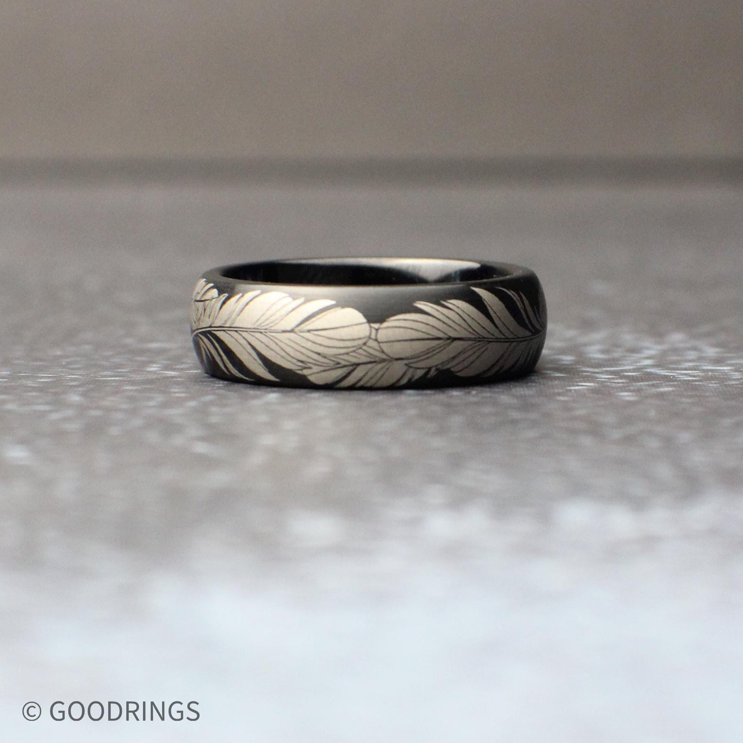 6mm black tungsten ring with a continuous silver feather engraving wrapped fully around the band, shown resting on a textured gray surface with a clean, minimal background.