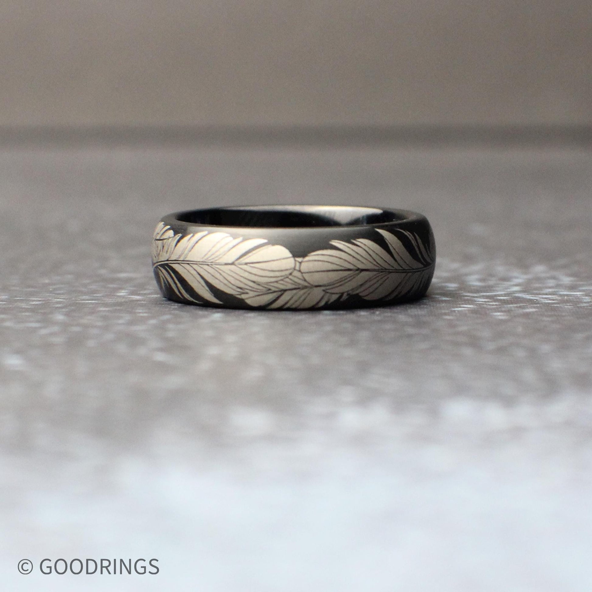 6mm black tungsten ring with a continuous silver feather engraving wrapped fully around the band, shown resting on a textured gray surface with a clean, minimal background.