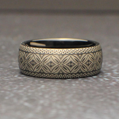 Close up of a black tungsten ring featuring a continuous Polynesian tribal engraving pattern wrapping around the entire band. The silver gray etched design contrasts against the dark metal creating a bold mens wedding band photographed on natural wood.