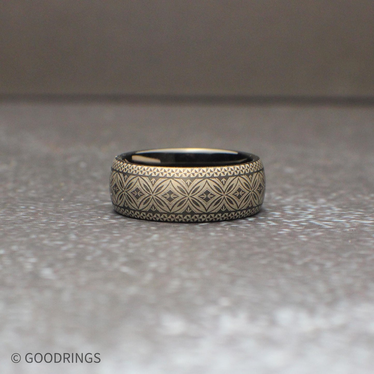 Close up of a black tungsten ring featuring a continuous Polynesian tribal engraving pattern wrapping around the entire band. The silver gray etched design contrasts against the dark metal creating a bold mens wedding band photographed on natural wood.