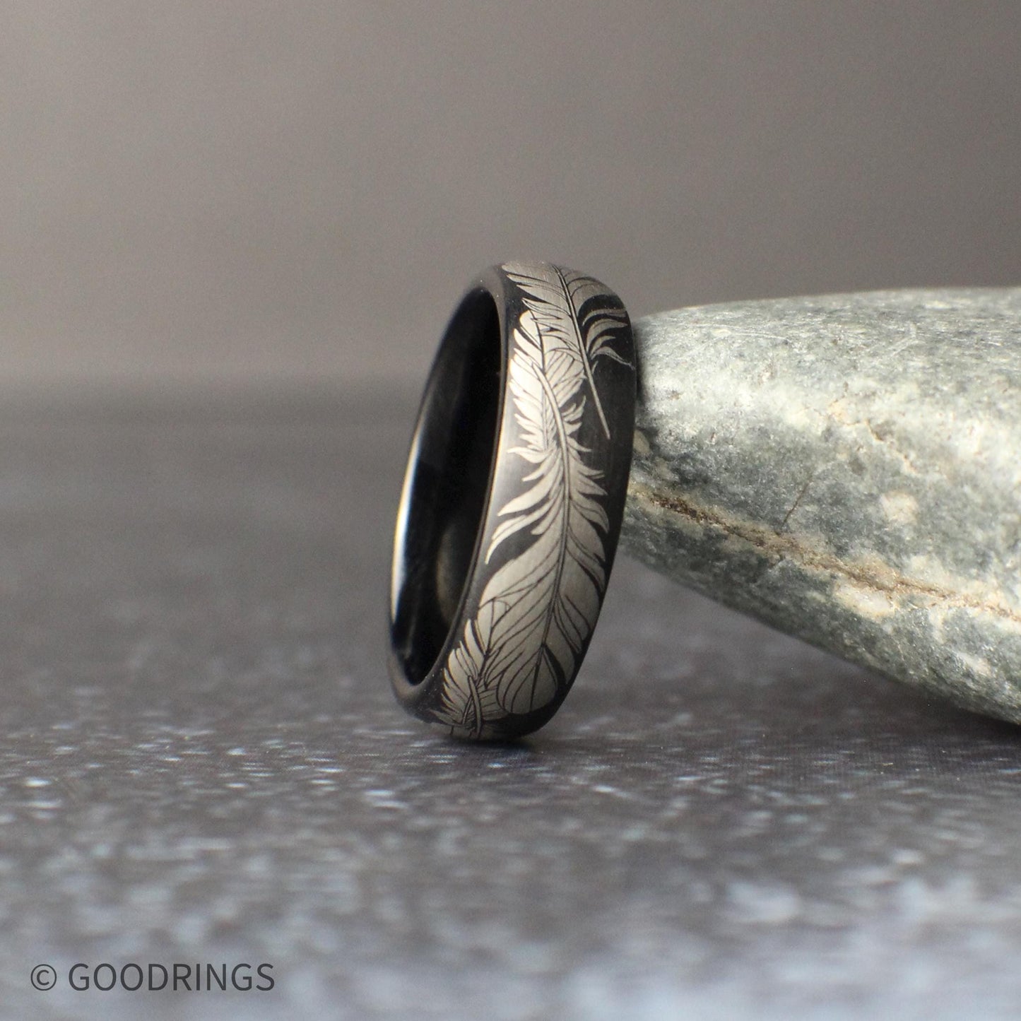 Close-up side angle of a black tungsten feather ring leaning against a smooth stone, highlighting the detailed silver engraved feather pattern and polished comfort fit interior.