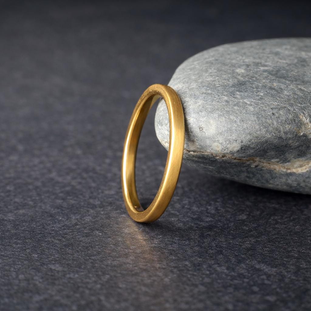Gold tungsten wedding band leaning against a smooth gray stone on a dark textured background, featuring a brushed finish and polished inner band. The composition highlights contrast between the warm gold ring and cool stone, creating a natural and modern aesthetic. Ideal minimalist wedding band for men or women with a comfort fit design.