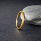 Gold tungsten wedding band leaning against a smooth gray stone on a dark textured background, featuring a brushed finish and polished inner band. The composition highlights contrast between the warm gold ring and cool stone, creating a natural and modern aesthetic. Ideal minimalist wedding band for men or women with a comfort fit design.