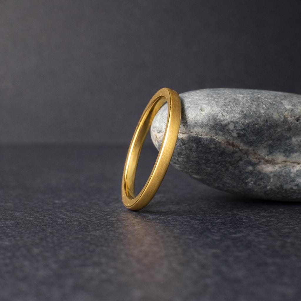 Minimalist gold tungsten ring angled and leaning against a rounded gray stone, set on a dark textured surface with soft lighting. The brushed matte finish contrasts with the polished interior, giving a refined and modern look. Perfect simple wedding band for men or women, showcasing durability, comfort fit, and timeless style.