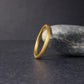 Minimalist gold tungsten ring angled and leaning against a rounded gray stone, set on a dark textured surface with soft lighting. The brushed matte finish contrasts with the polished interior, giving a refined and modern look. Perfect simple wedding band for men or women, showcasing durability, comfort fit, and timeless style.