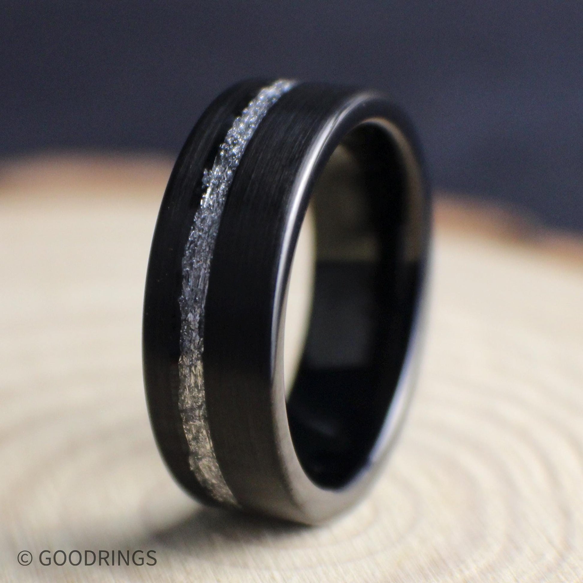 Men’s black tungsten ring featuring a brushed finish and offset silver leaf inlay, displayed upright on natural wood.