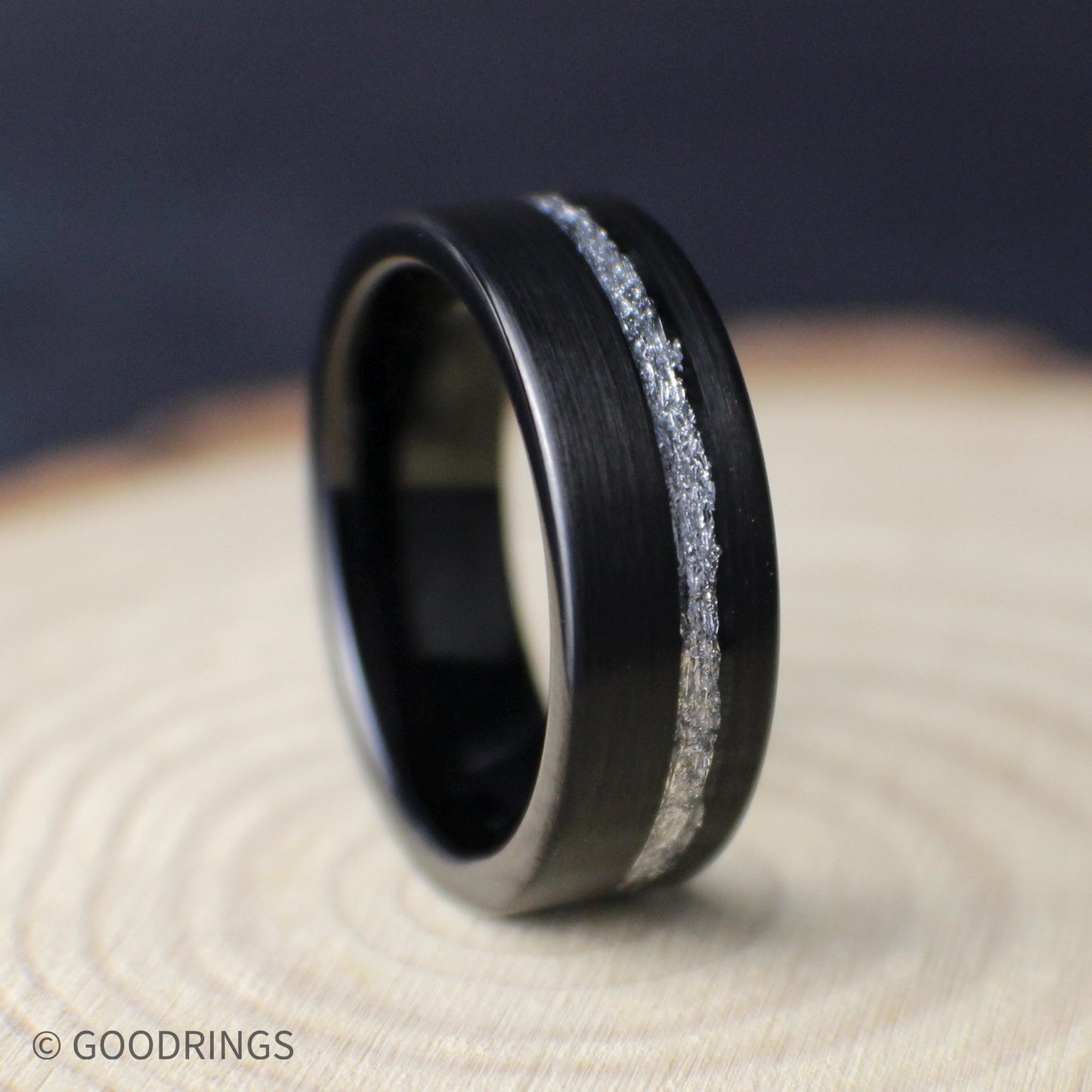 Black tungsten men’s wedding ring with brushed finish and offset genuine silver leaf inlay, shown on a light wood background.
