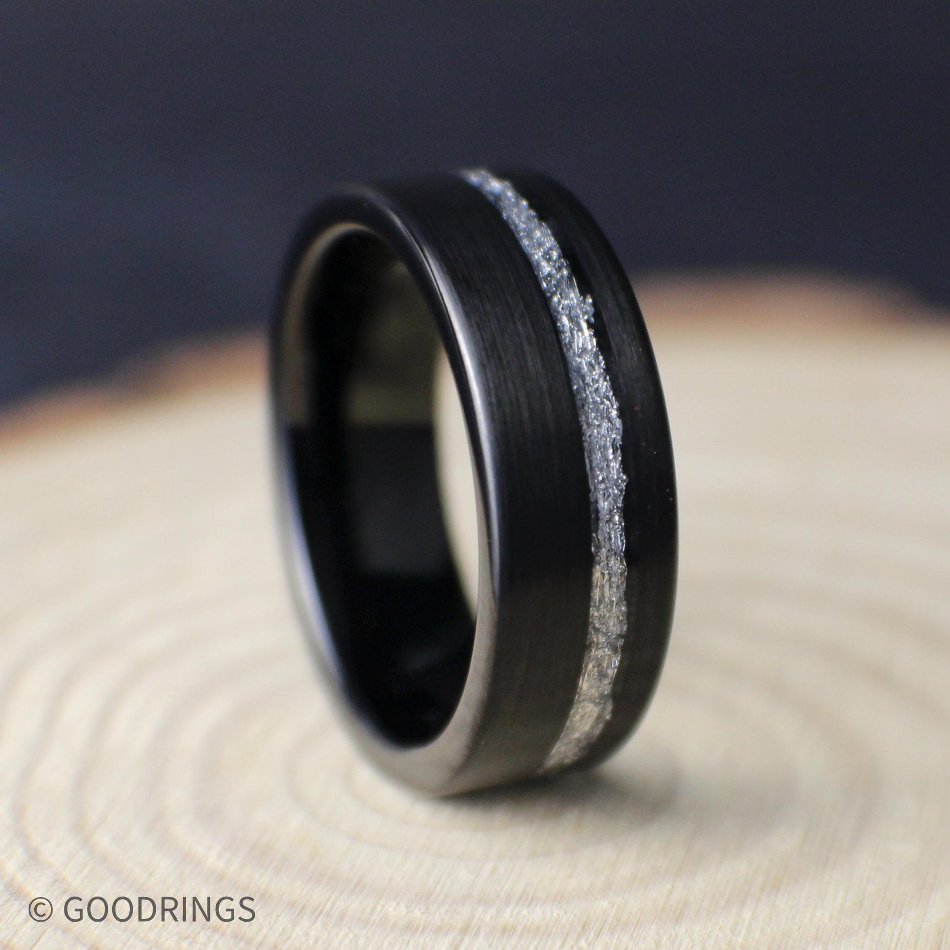 Black tungsten men’s wedding ring with brushed finish and offset genuine silver leaf inlay, shown on a light wood background.