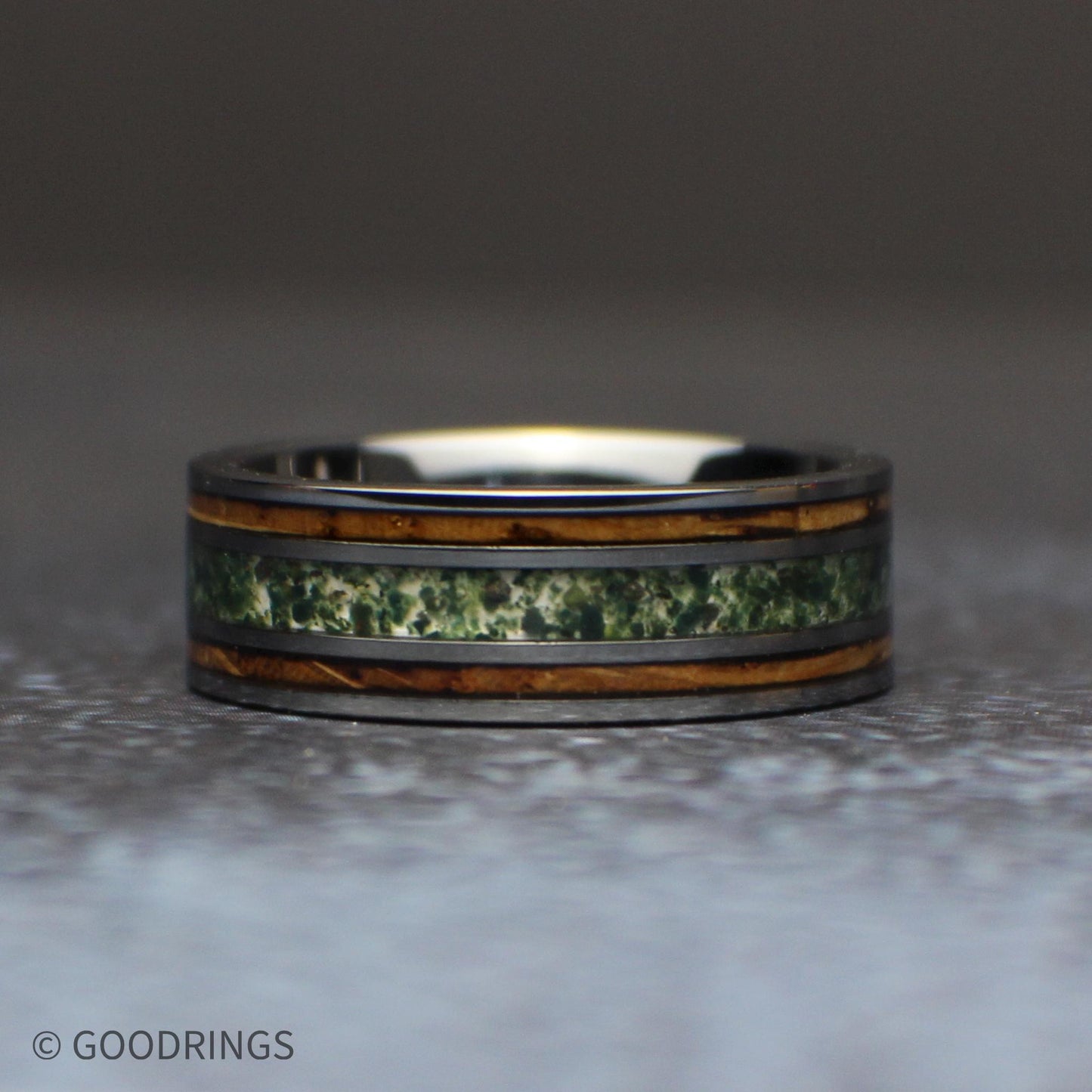 Angled view of an 8mm gunmetal tungsten ring standing on a light wood block, showing the green moss agate center and two brown whiskey barrel wood inlays.
