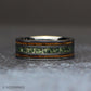 Angled view of an 8mm gunmetal tungsten ring standing on a light wood block, showing the green moss agate center and two brown whiskey barrel wood inlays.