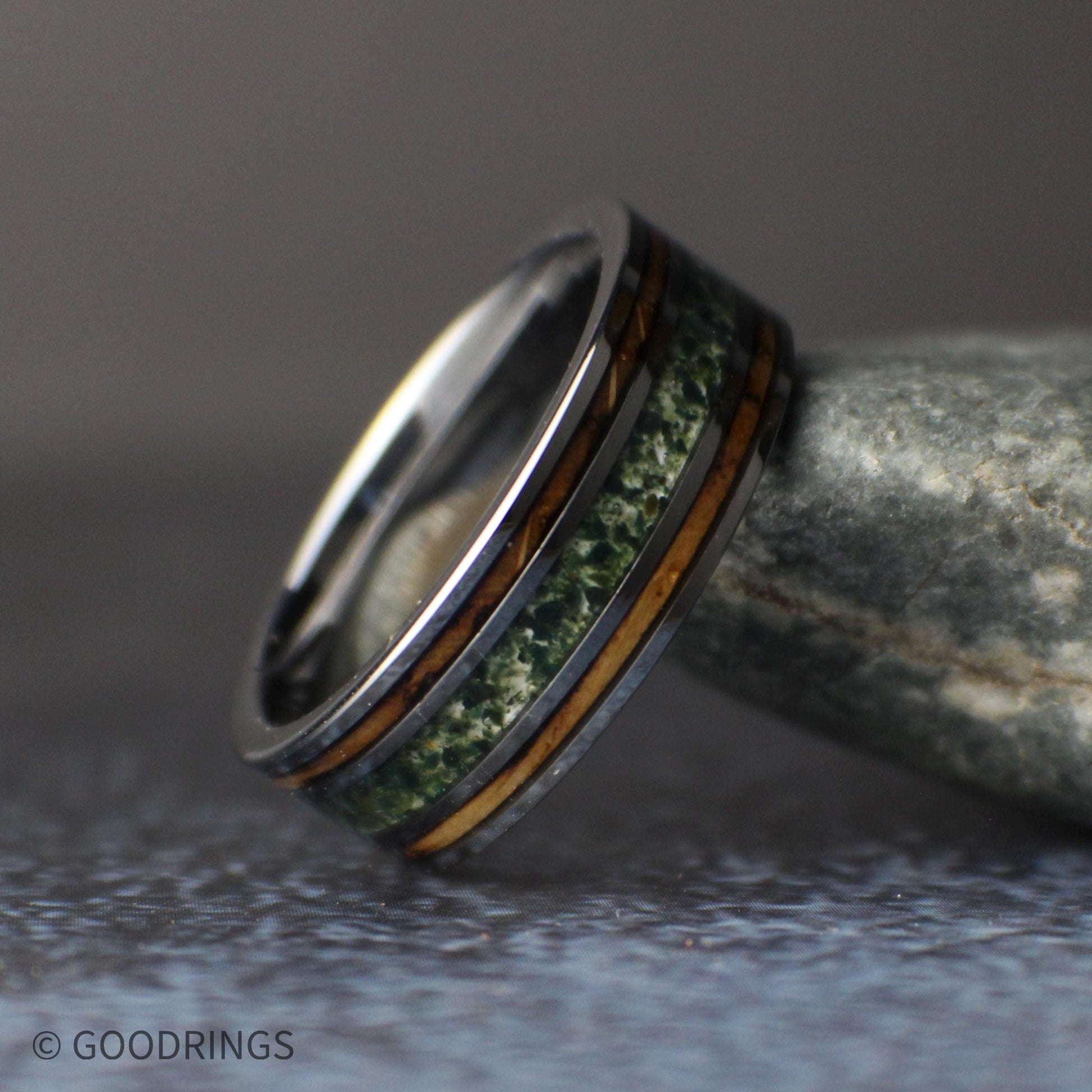 Front view of a men’s gunmetal tungsten wedding band standing upright, displaying its wide moss agate center inlay and two narrow whiskey barrel wood stripes.